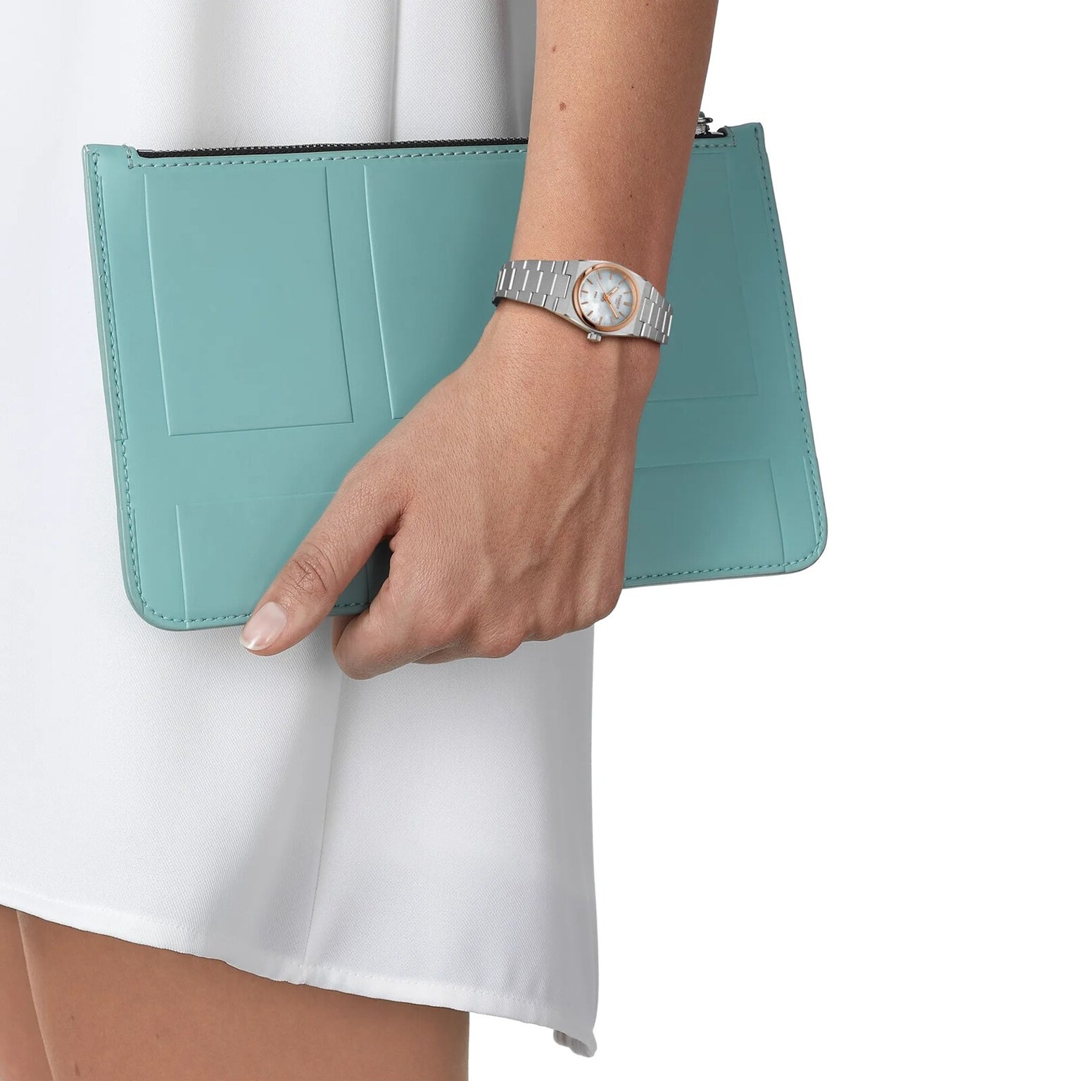 A person wearing a Tissot PRX 25mm silver wristwatch with sapphire crystal holds a light blue clutch purse, shown against a white outfit and plain background.