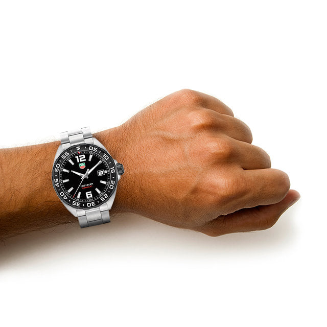 A wrist dons the TAG Heuer Formula 1 Date watch, featuring a stainless steel case, black round face, white hour markers, and a rotating bezel, shown against a plain white background.