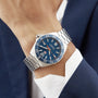 A person wearing a TAG Heuer Formula 1 Date watch with a blue sunray dial, white markers, and stainless steel band, dressed in a dark blue suit and white shirt.