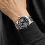 A person wearing a TAG Heuer Formula 1 Date silver metal wristwatch with a black dial, bold white numbers, and tachymeter bezel, dressed in a black long-sleeve top.