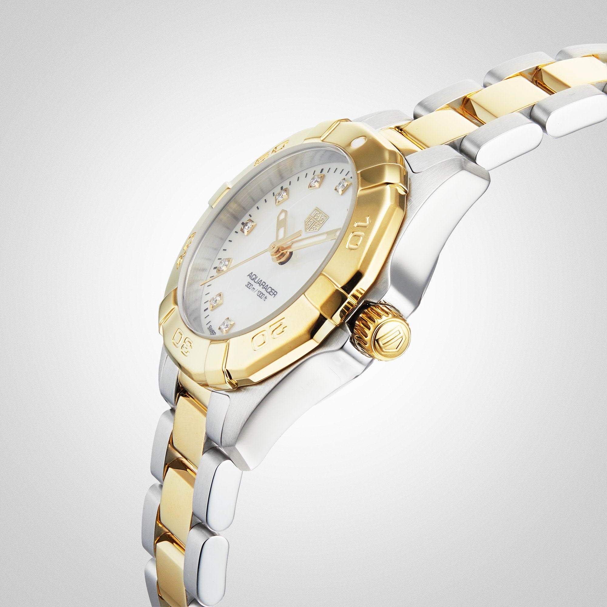 The TAG Heuer Aquaracer Date, a two-tone 18K gold and stainless steel wristwatch with white dial and gold bezel, is displayed at an angle on a light gradient background.