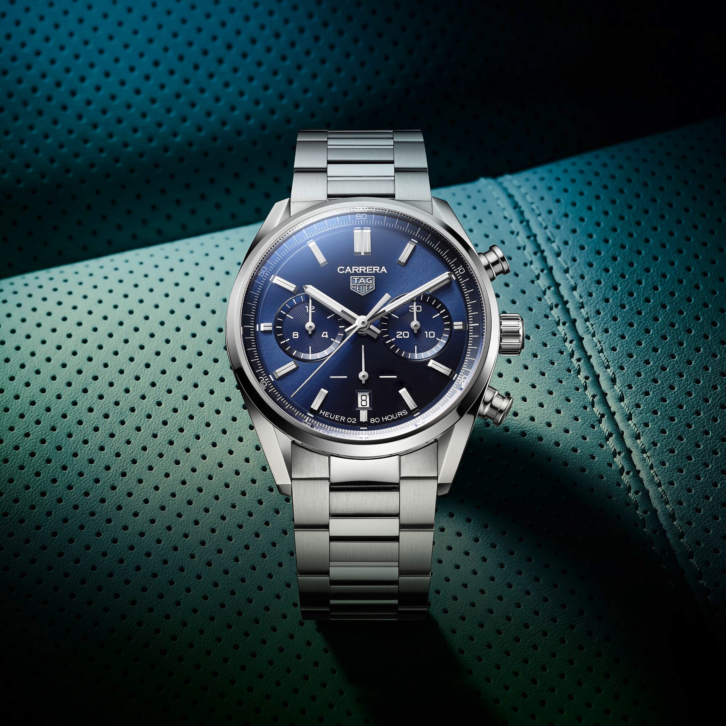 The TAG Heuer Carrera Chronograph, featuring a blue dial, chronograph subdials, and date display, is set on a textured teal surface, highlighting its motor-racing heritage.