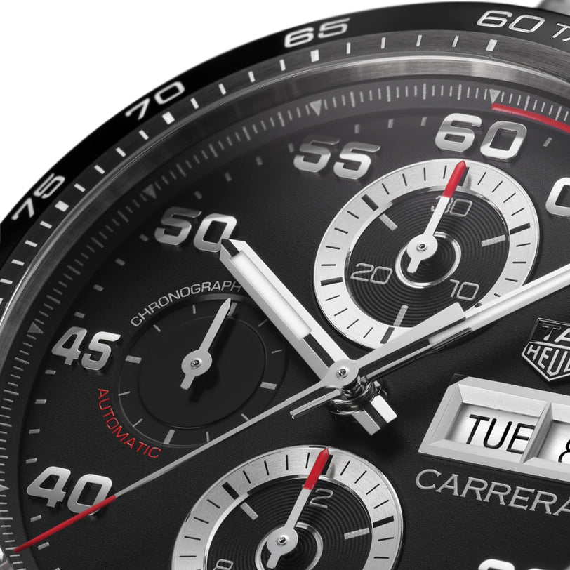 Close-up of the TAG Heuer Carrera Chronograph watch face, featuring a black ceramic bezel, three subdials, white hands, and a day-date window showing TUE 8.