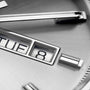 Close-up of the TAG Heuer Carrera Day-Date watch face, showing metallic hour markers, calibre 5 movement, and a partial day-date window with TUE 8 on a brushed silver background.