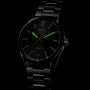 The TAG Heuer Carrera Date watch features a black dial, steel case, and metal band. In low light, its green luminescent hour markers and hands stand out. Powered by the precision Calibre 5 movement.