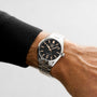 A person wearing a TAG Heuer Carrera Date watch with a steel case, silver stainless steel band, black dial, and date display on their left wrist.
