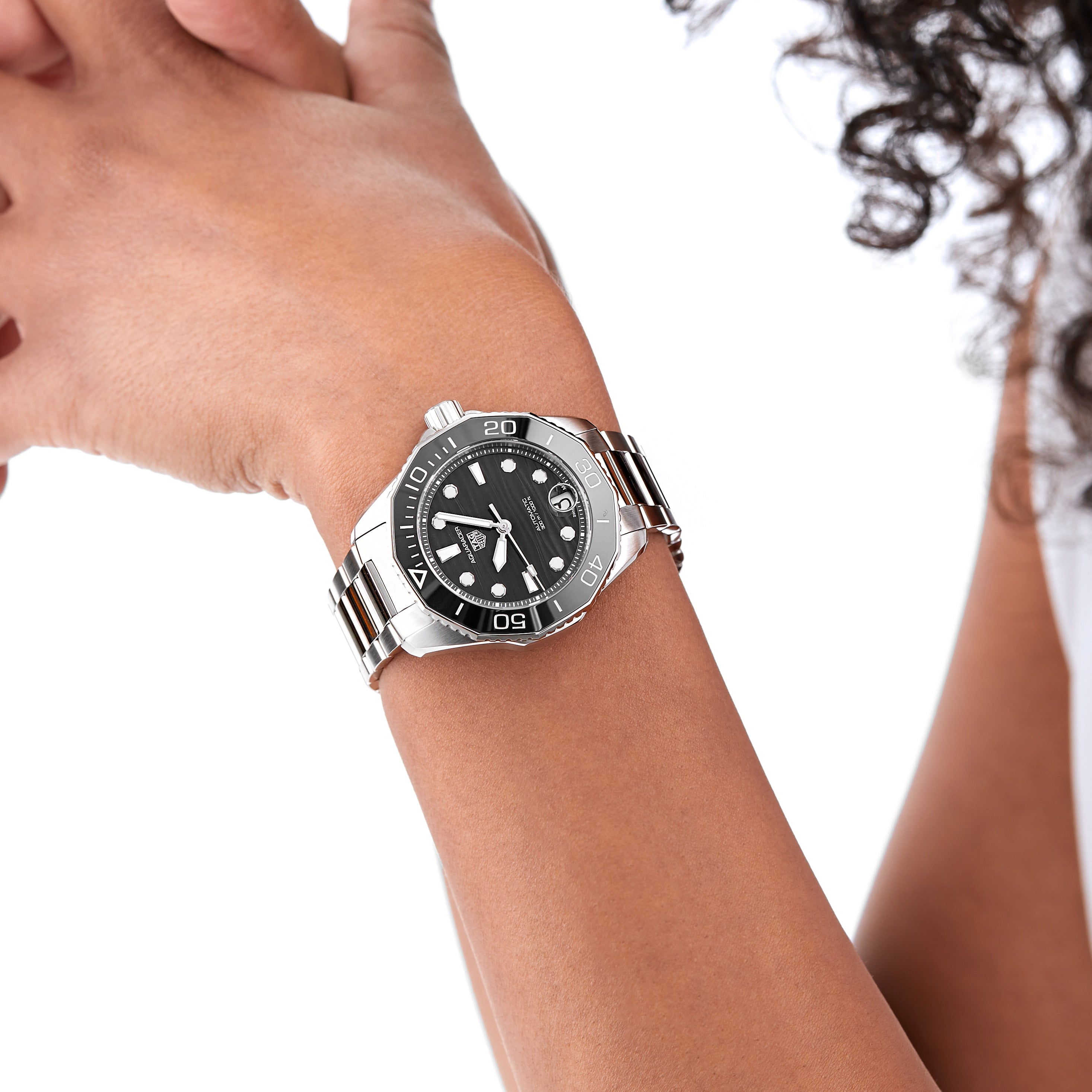 A person wearing a TAG Heuer Aquaracer Professional 300 Date with a stainless steel case and black dial holds their arm near their face against a white background.