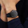 A person with arms crossed against a dark background wears a TAG Heuer Connected Calibre E4 Porsche Edition on their left wrist, featuring a black and blue strap and an inactive watch face.