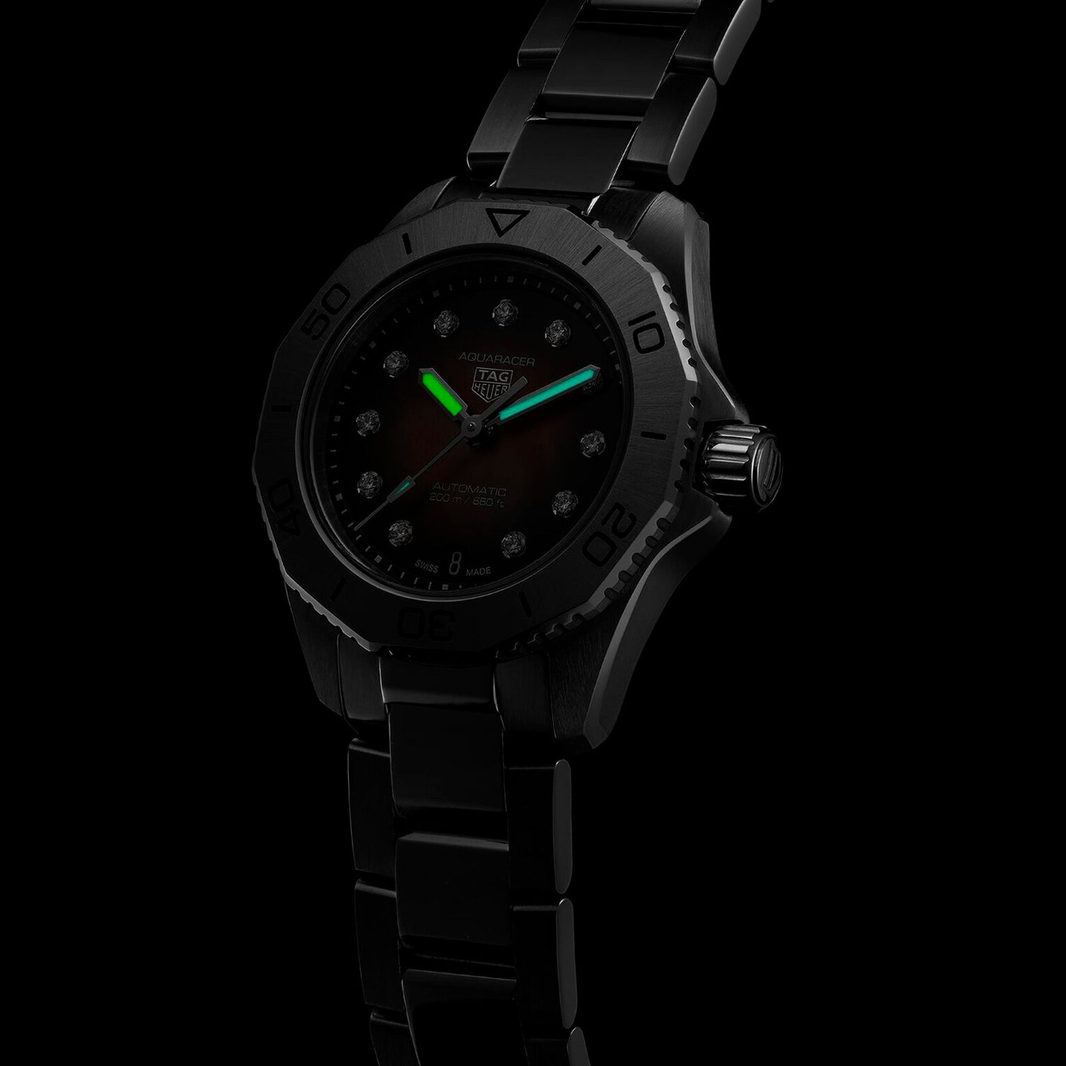 A TAG Heuer Aquaracer Professional 200 Date wristwatch with a stainless steel case and metal band glows in low light, showcasing its luminous green hour and minute hands.