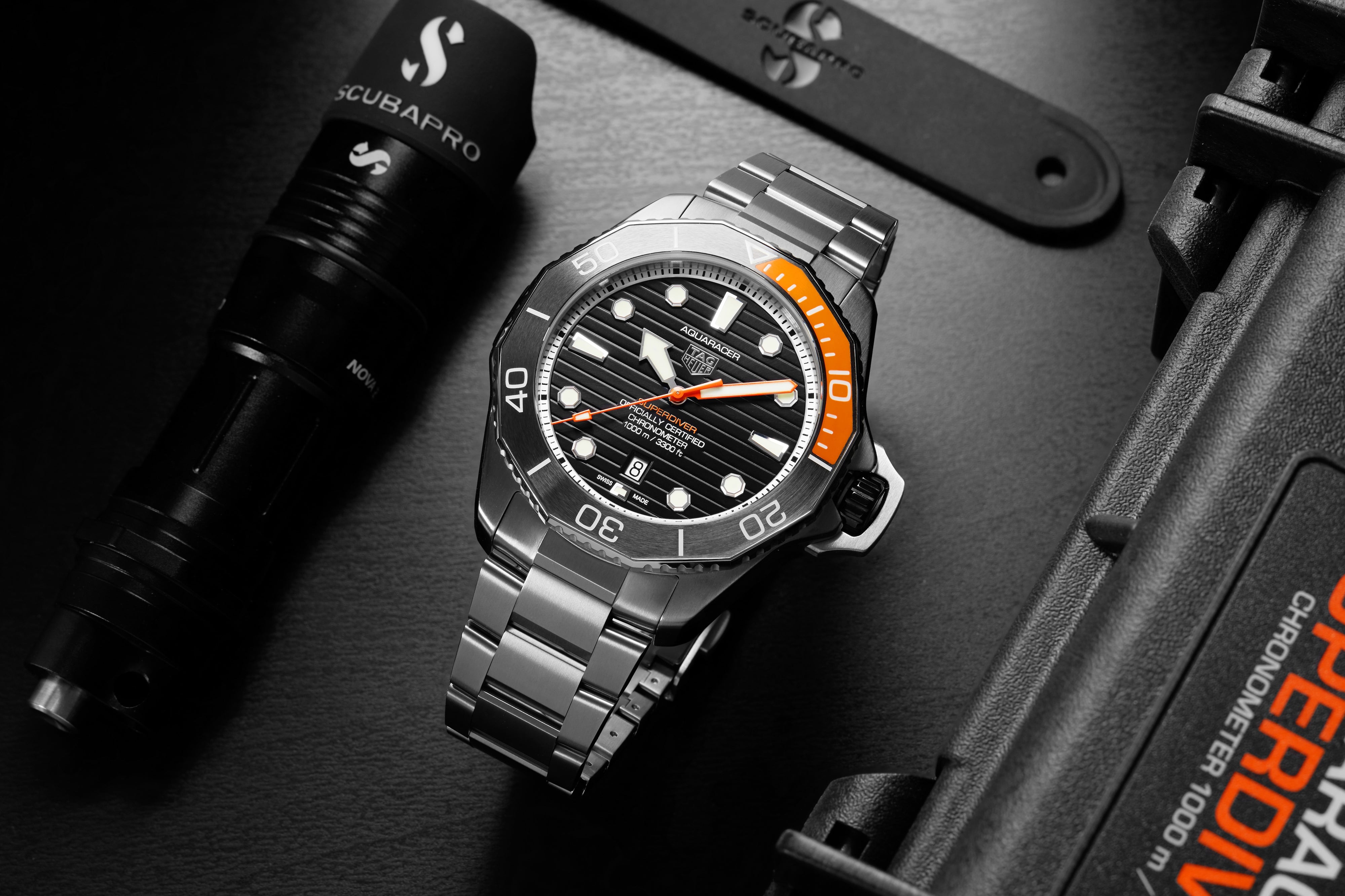 The TAG Heuer Aquaracer Professional 1000 Superdiver, featuring a stainless steel case and a black and orange bezel, is shown beside scuba gear and a flashlight, emphasizing its outstanding water resistance.