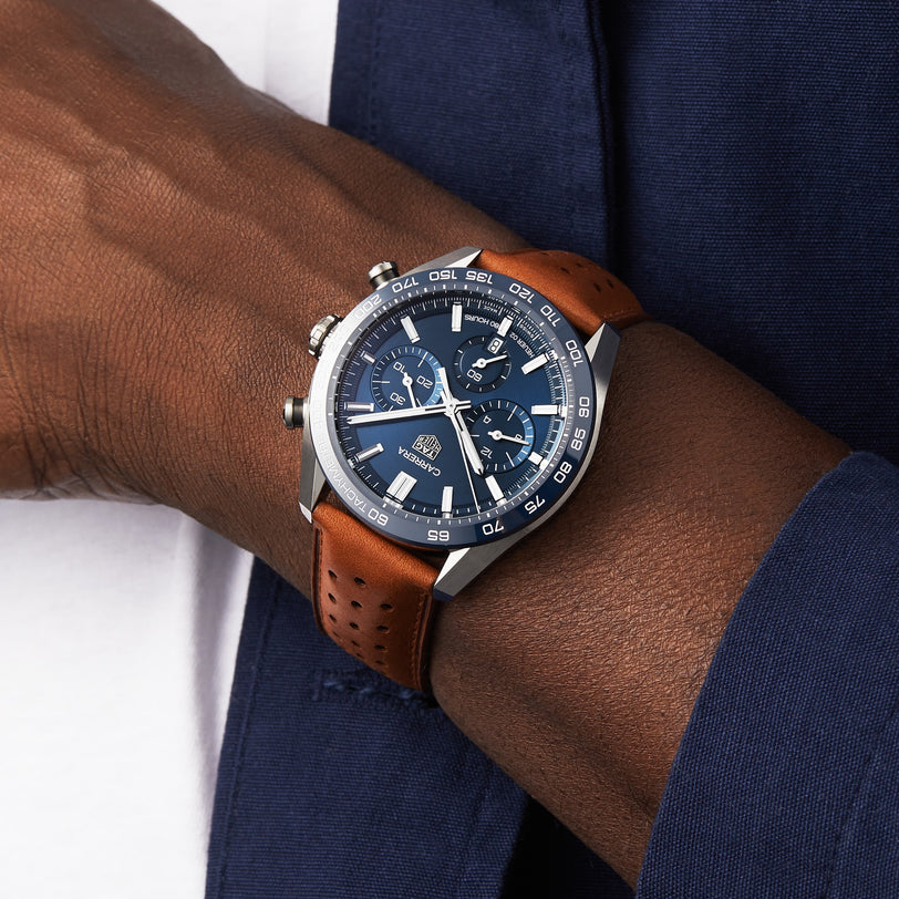 A TAG Heuer Carrera Chronograph with a brown leather strap and black dial is worn on a wrist beside a navy blue sleeve.