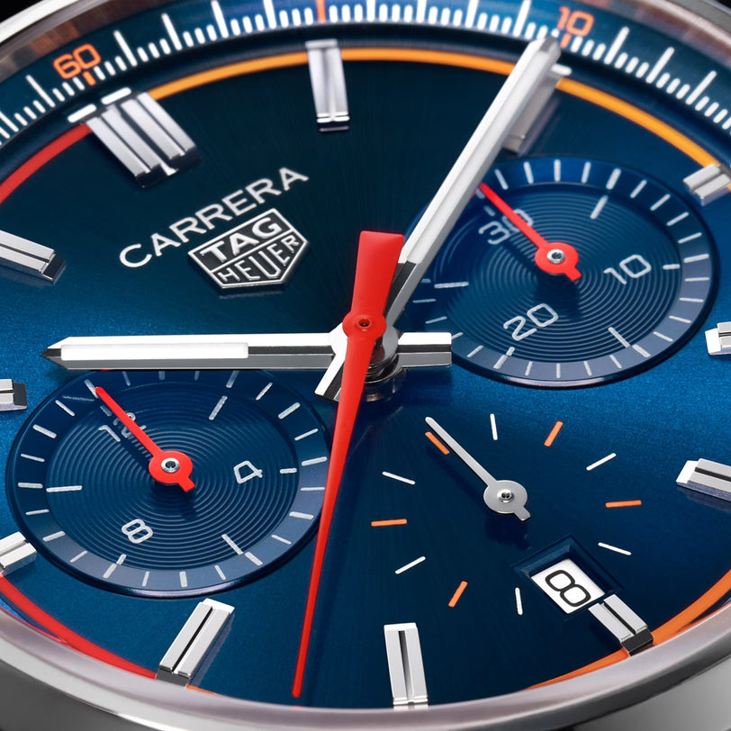 Close-up of a blue TAG Heuer Carrera Chronograph watch face, featuring red and white hands, two subdials, and a date window showing the number 8.