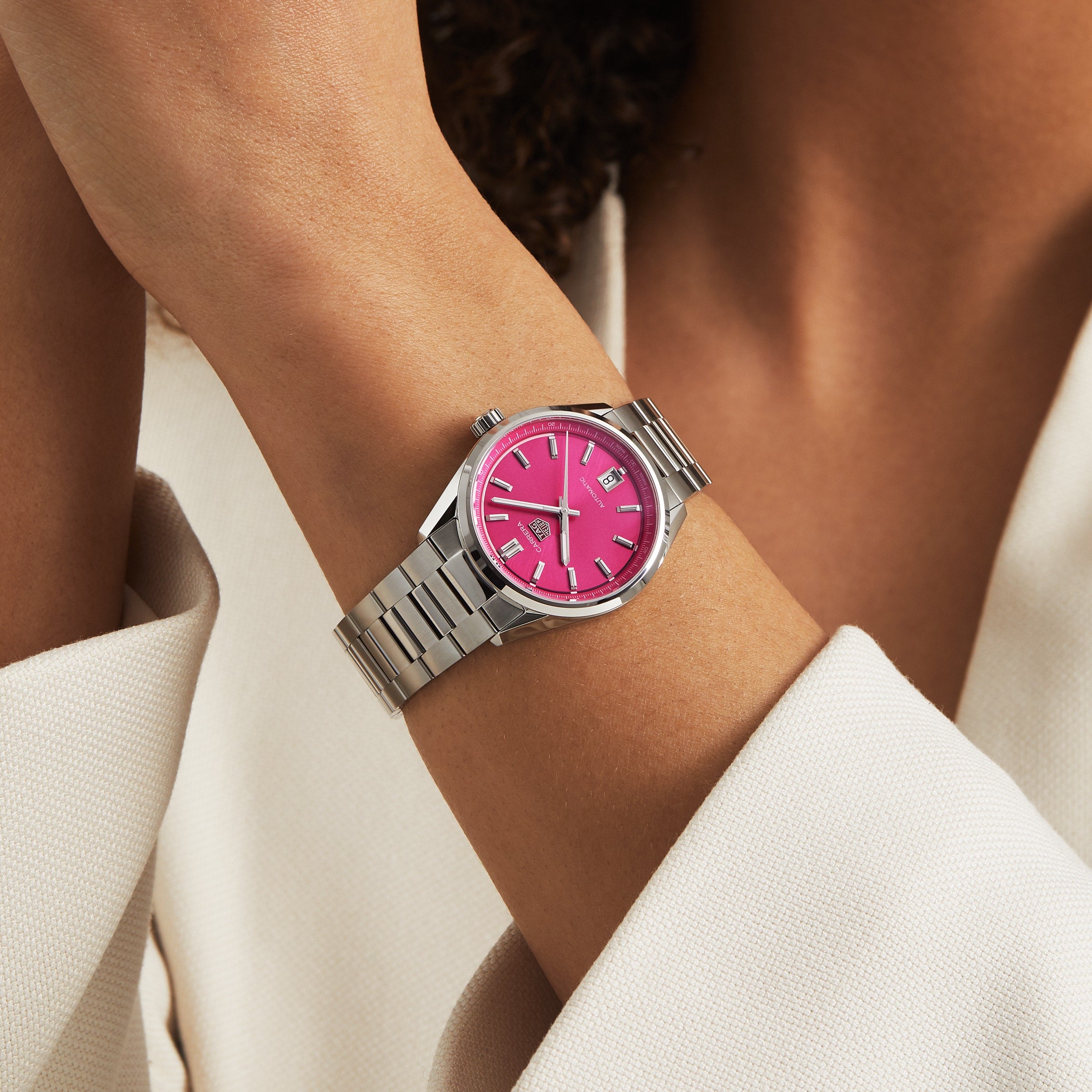 Wearing a TAG Heuer Carrera Date with a silver case, bright pink dial, and silver hour markers, paired with a light blazer—a stylish ensemble that blends elegance and endurance.