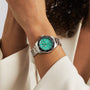 A person with curly hair, dressed in a cream-colored jacket, wears a TAG Heuer Aquaracer Professional 300 Date sports watch with a green dial and black bezel.