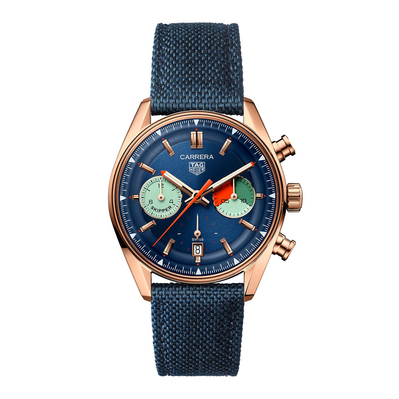 The TAG Heuer Carrera Chronograph Skipper features a gold case, blue textured strap, blue dial with two green subdials, orange chronograph hand, chronograph movement, and a date display at 6 o’clock.