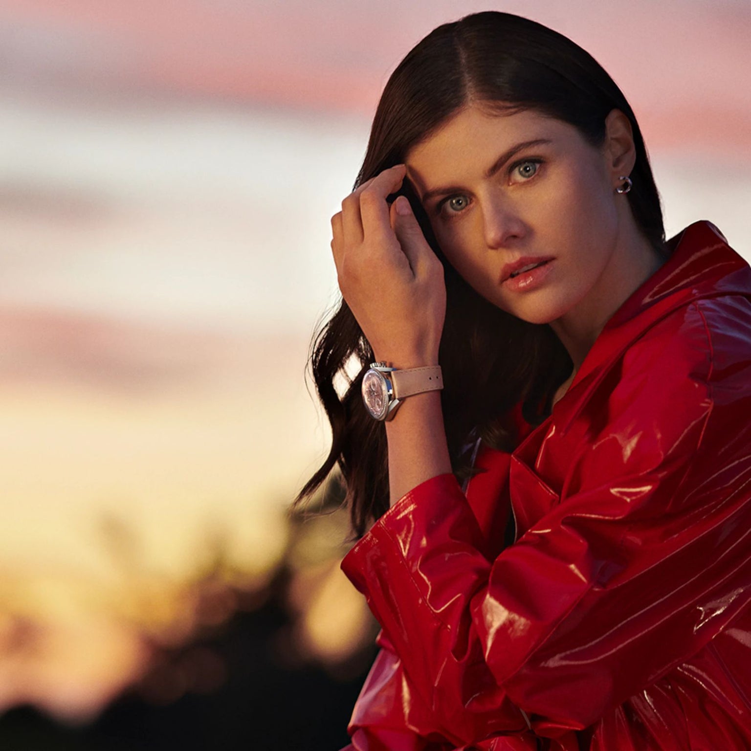 Woman in a shiny red jacket poses outdoors at sunset, touching her hair and wearing a TAG Heuer Carrera Chronograph wristwatch.