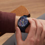 A person wearing the TAG Heuer Limited Edition Formula 1 Chronograph X Oracle Red Bull Racing with a vibrant dial adjusts the bezel while seated on a wooden chair.