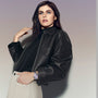 Woman with long dark hair in a black leather jacket and beige pants, glancing upward, showcases the TAG Heuer Carrera Date watch.