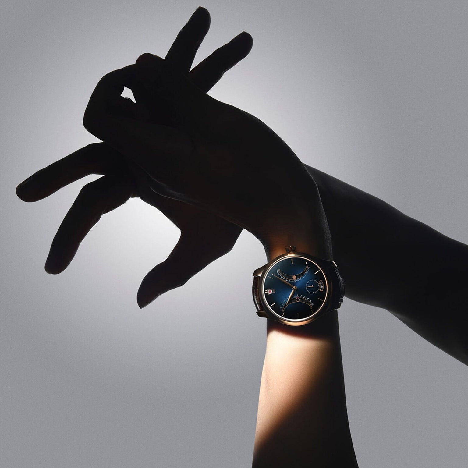 A person’s hand wearing the H. Moser & Cie Endeavour Chinese Calendar with a blue face, posed artistically against a gray background.