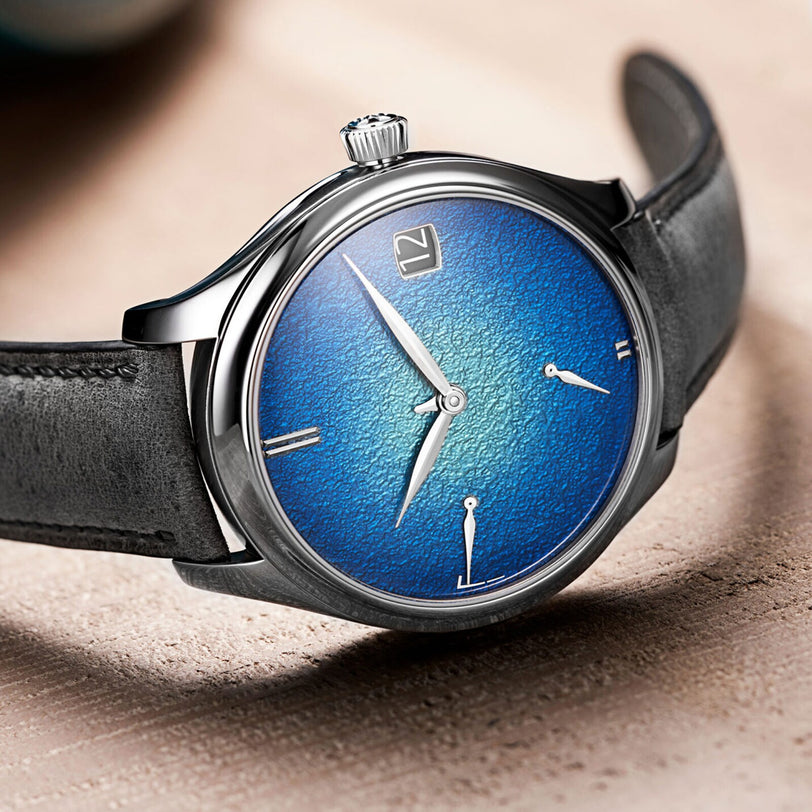 The H. Moser & Cie Endeavour Perpetual Calendar features a textured blue grand feu enamel dial, silver hour and minute hands, minimal hour markers, and a black leather strap, shown resting on a light surface.