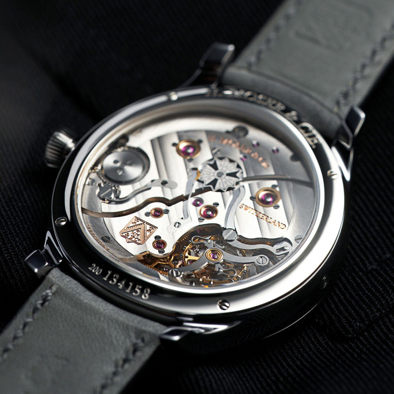 Close-up of the H. Moser & Cie Endeavour Perpetual Calendar’s back, revealing its mechanical movement through a transparent case, with visible gears, jewels, engravings, and exquisite grand feu enamel.
