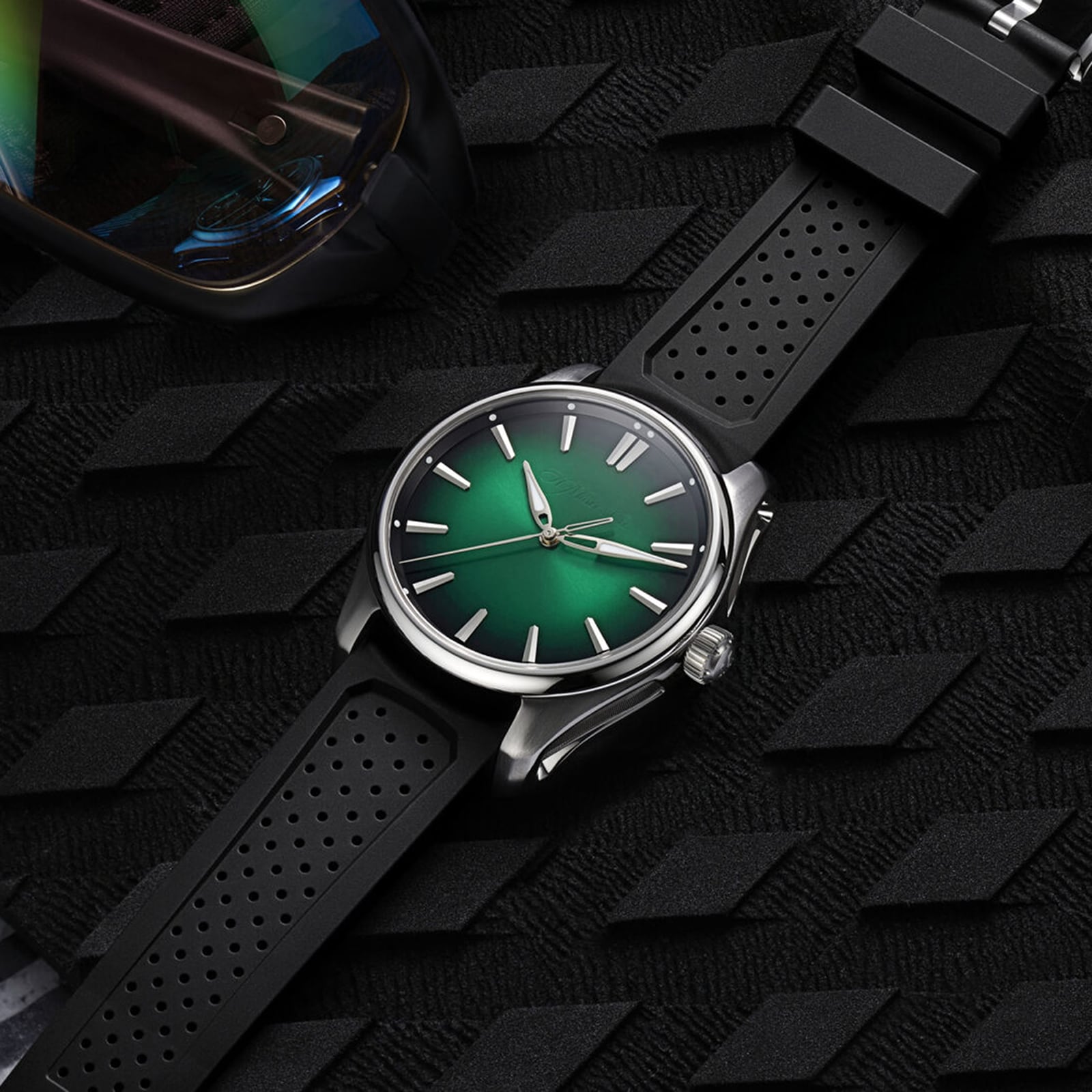 The H. Moser & Cie Pioneer Centre Seconds Cosmic Green wristwatch with a green dial and black strap rests on a textured black surface next to a pair of sunglasses.