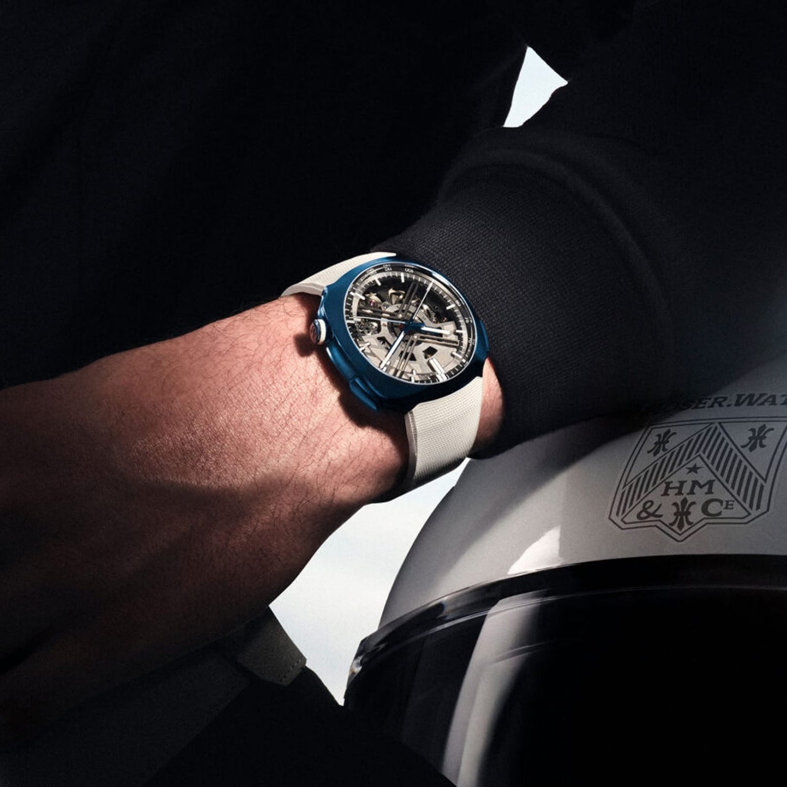 A person wearing the H. Moser & Cie Streamliner Alpine Divers Edition wristwatch, featuring a blue and white design, rests their hand on a helmet with a crest logo.