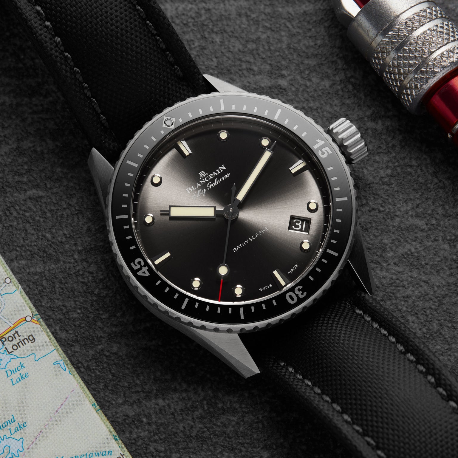 A close-up of a Blancpain Fifty Fathoms Bathyscaphe diver’s watch with a black dial, black strap, and date window, featuring an automatic movement, placed on a surface beside part of a map and a metal tool.