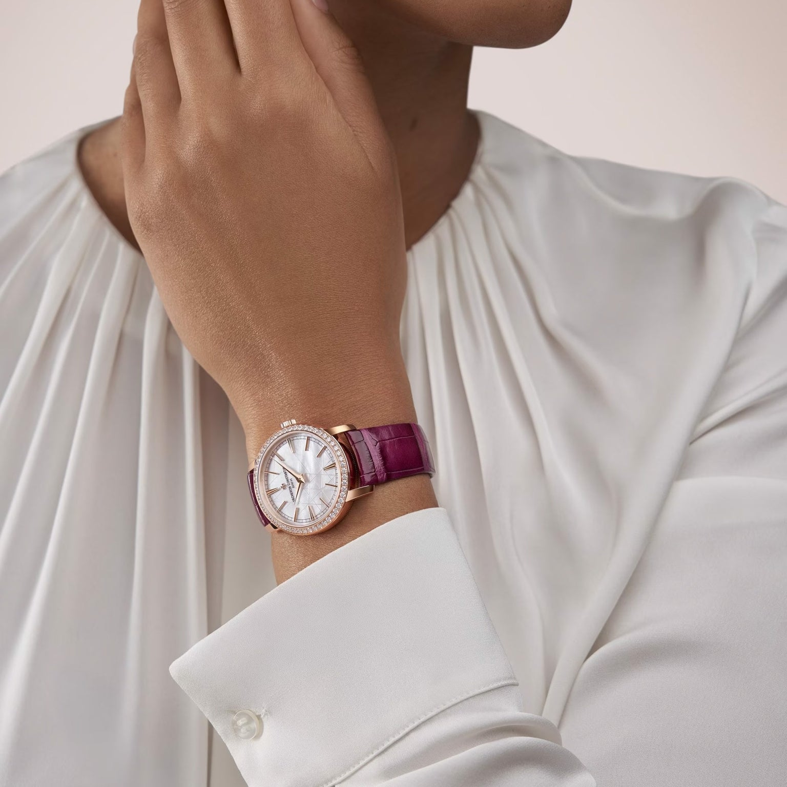 A person in a white blouse rests their hand on their face, showcasing a Vacheron Constantin Traditionnelle Manual-winding wristwatch with a pink leather strap and a round, jeweled dial.