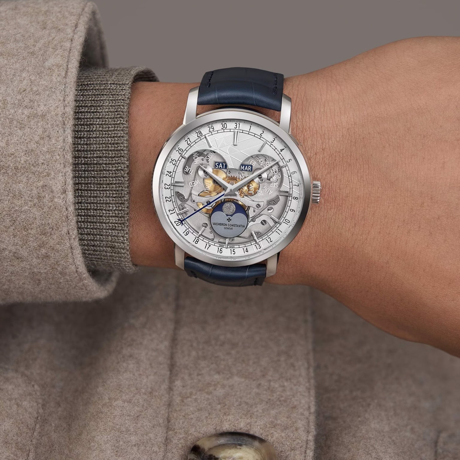 Wearing a beige coat, a person showcases the Vacheron Constantin Traditionnelle Complete Calendar Openface— a limited edition platinum watch with blue leather strap, exposed gears, and calendar displaying day, date, and moon phase.