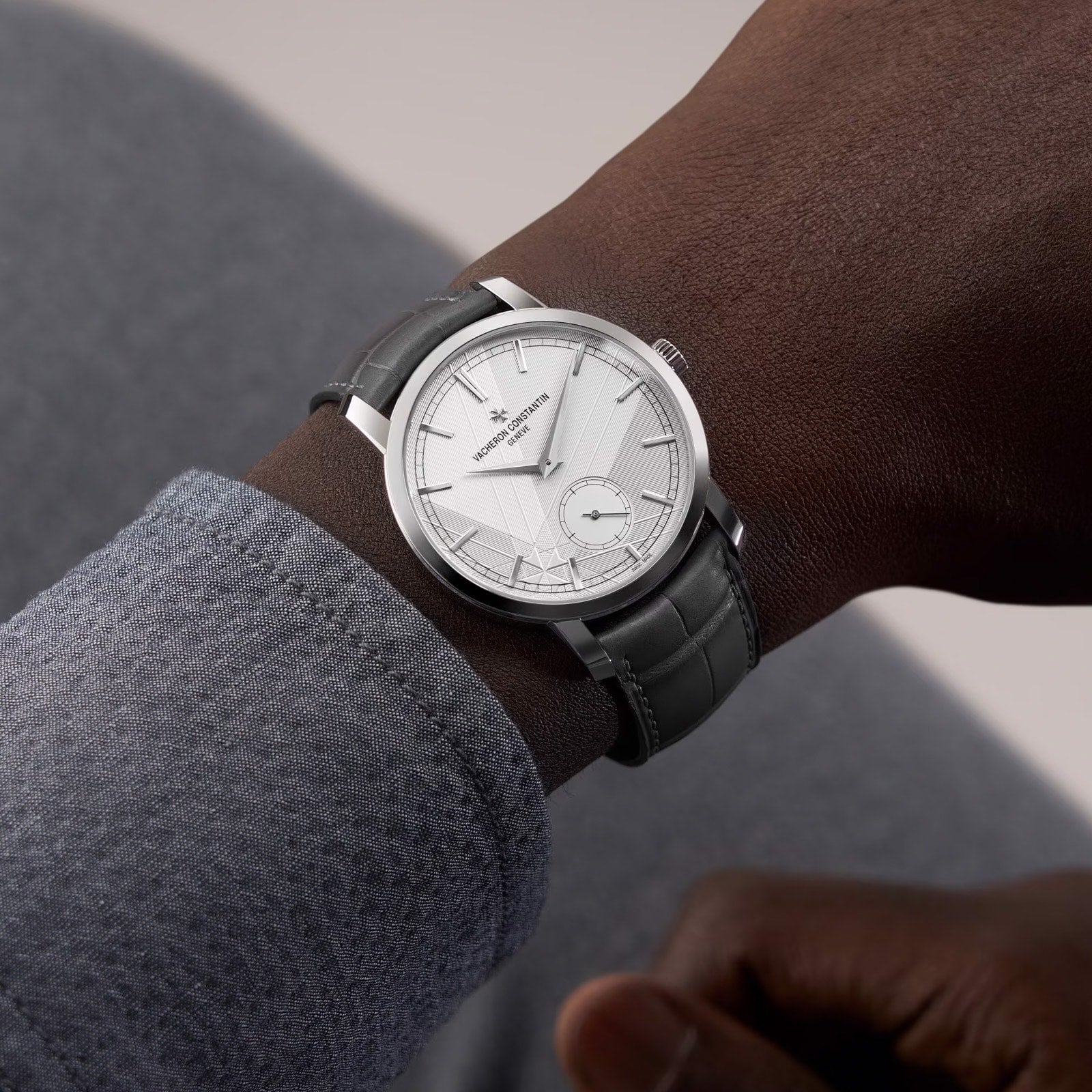 A close-up of a person wearing a Vacheron Constantin Traditionnelle Manual-Winding wristwatch featuring a white dial and gray leather strap.
