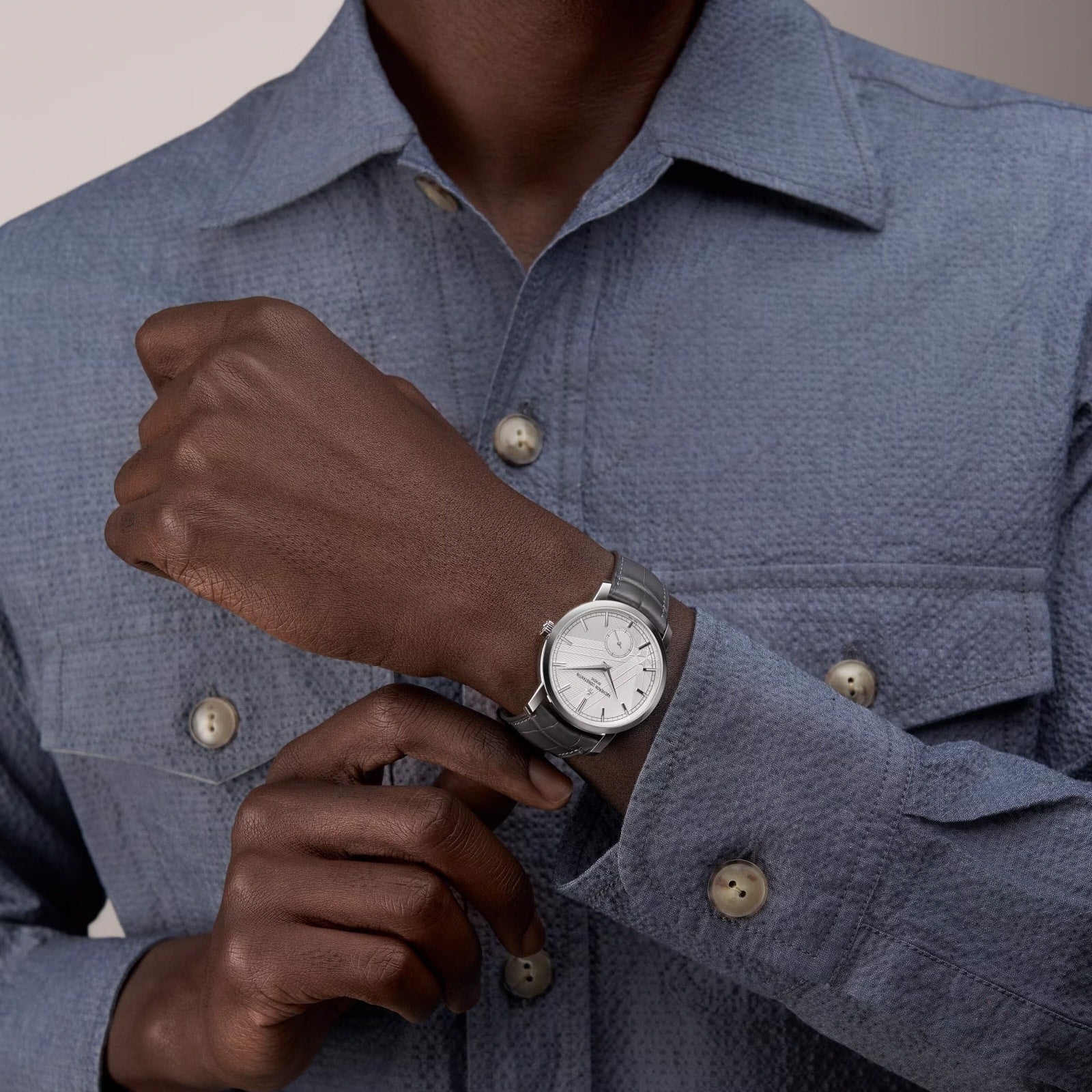 A person in a textured blue-gray button-up shirt showcases a Vacheron Constantin Traditionnelle Manual-Winding platinum watch with a white face on their left wrist.