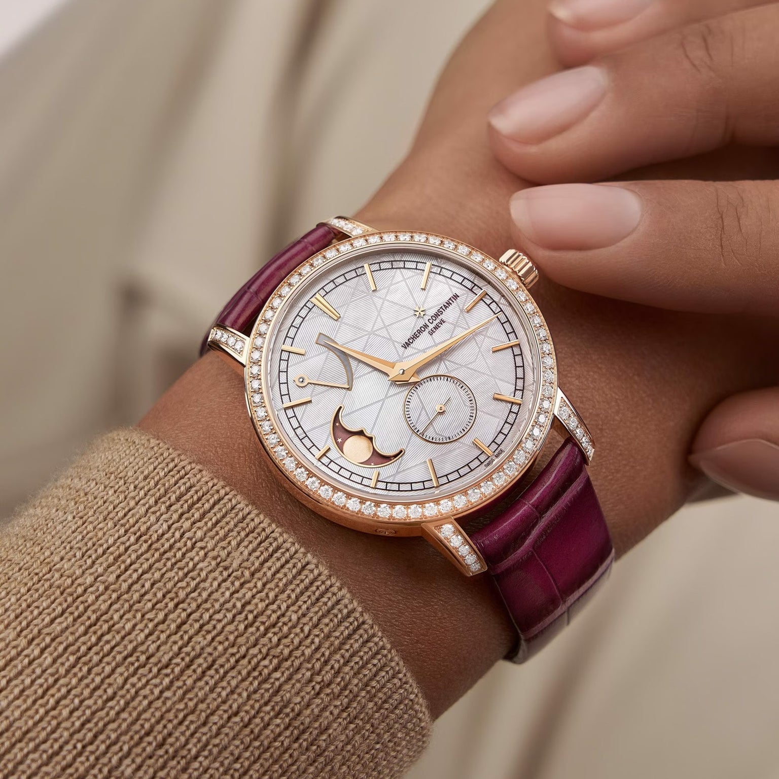 A close-up of a wrist wearing the Vacheron Constantin Traditionnelle Moon Phase in pink gold, featuring a diamond bezel, white dial with gold accents, moon phase display, and a burgundy leather strap.