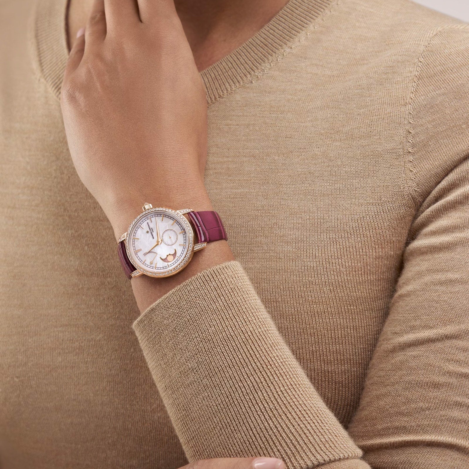 A person in a beige sweater rests their hand on their shoulder, showcasing the Vacheron Constantin Traditionnelle Moon Phase watch with pink gold, a burgundy leather strap, round white face, and an elegant moon phase detail.