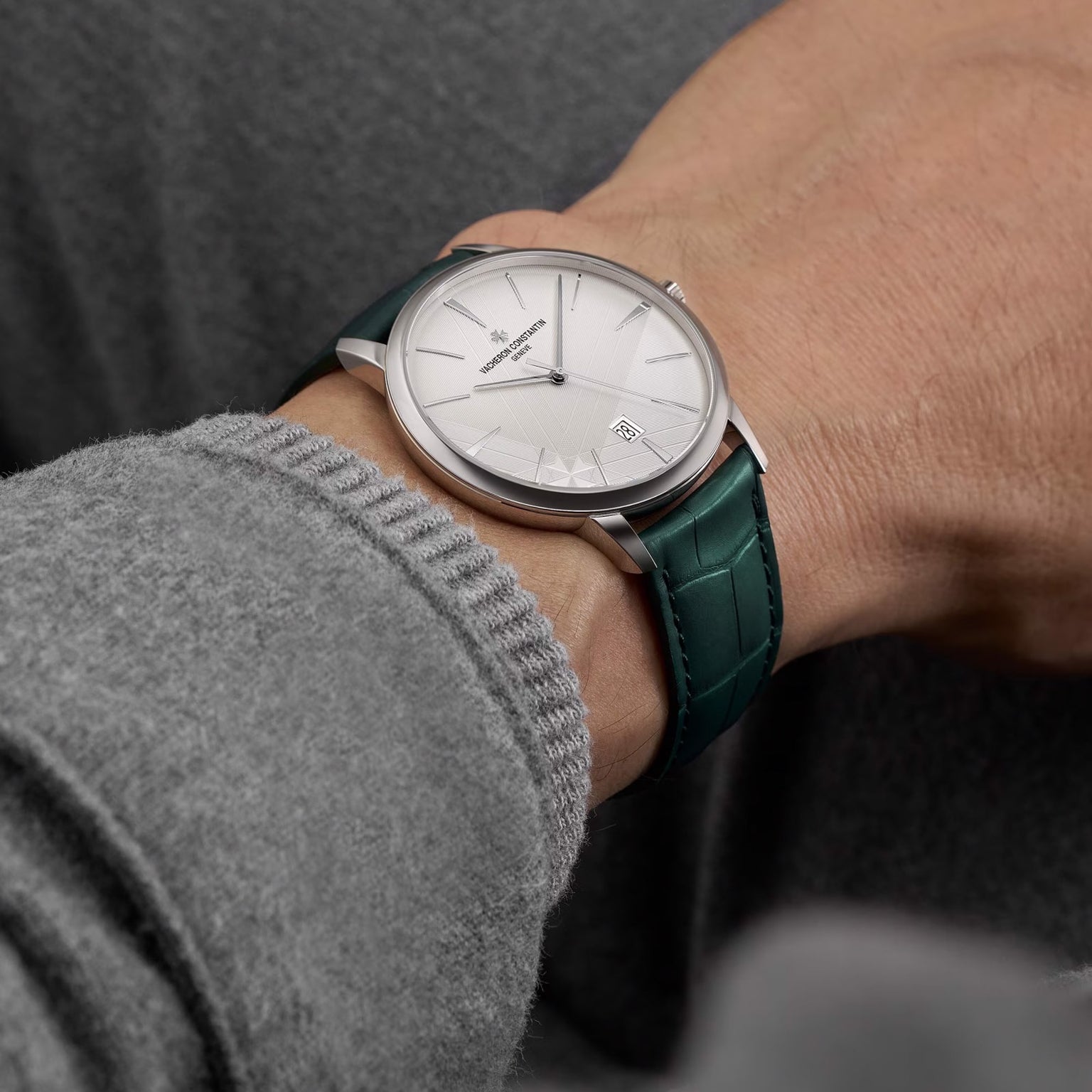 A person wearing the limited edition Vacheron Constantin Patrimony Self-Winding 18K white gold watch with a white dial and green leather strap, dressed in a grey long-sleeve garment.