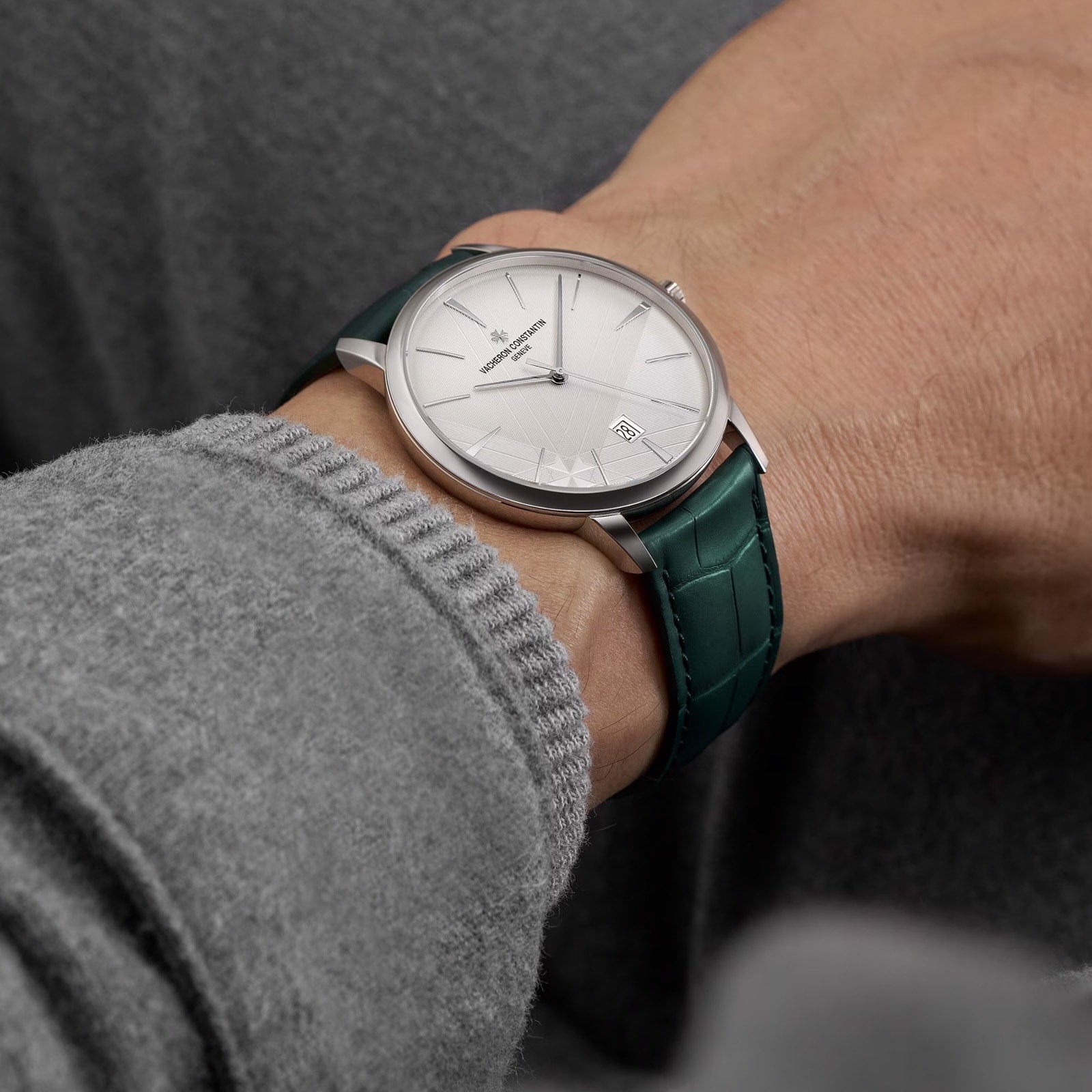 A person wearing the limited edition Vacheron Constantin Patrimony Self-Winding 18K white gold watch with a white dial and green leather strap, dressed in a grey long-sleeve garment.