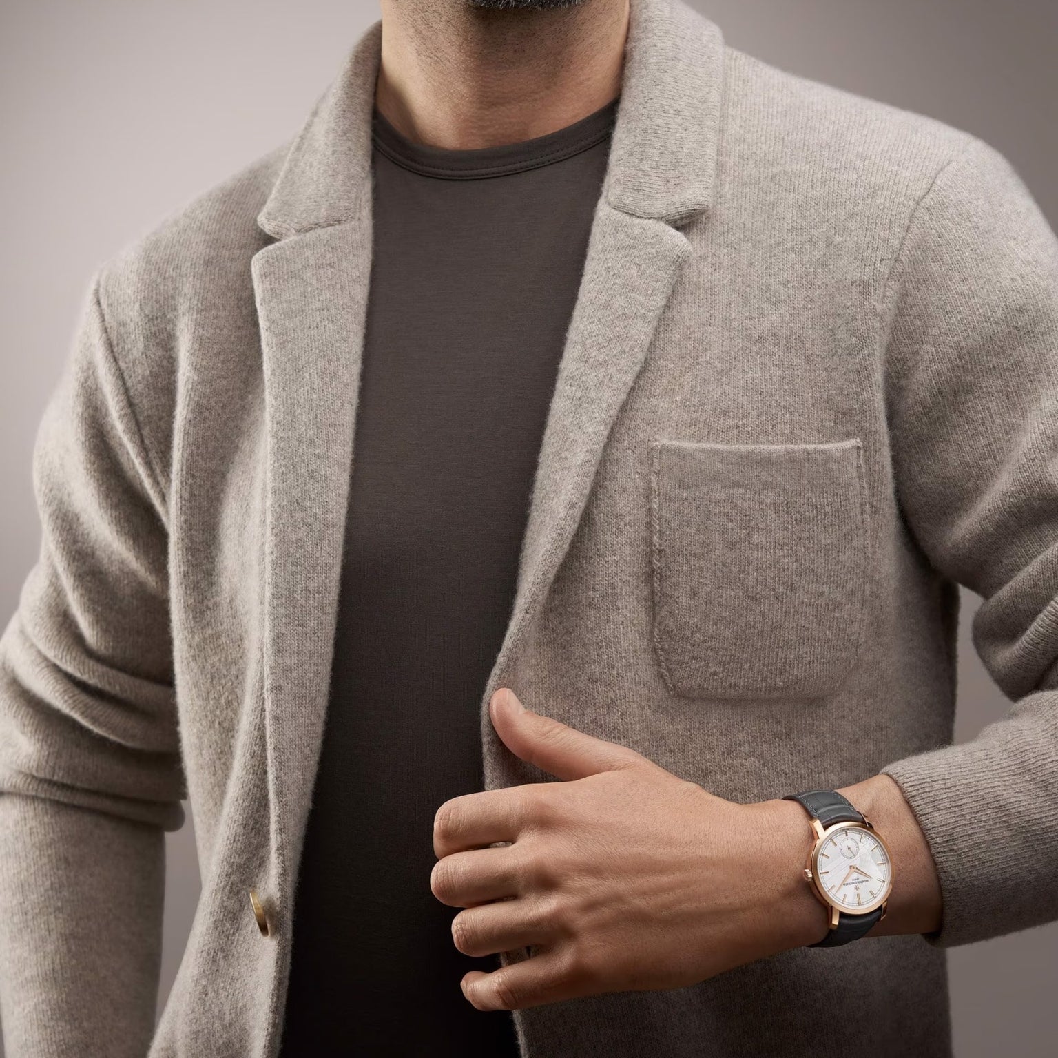 A person in a light gray blazer over a dark t-shirt displays a Vacheron Constantin Traditionnelle Manual-winding watch with a white face and black strap on their left wrist.