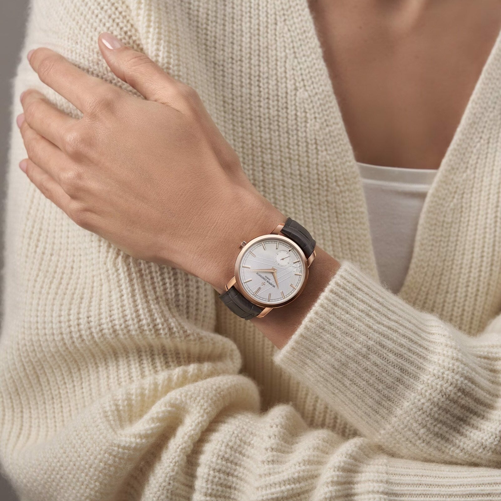 A person in a cream knit sweater crosses their arms, showcasing the limited edition Vacheron Constantin Traditionnelle Manual-winding watch with an 18K pink gold round case and black leather strap.