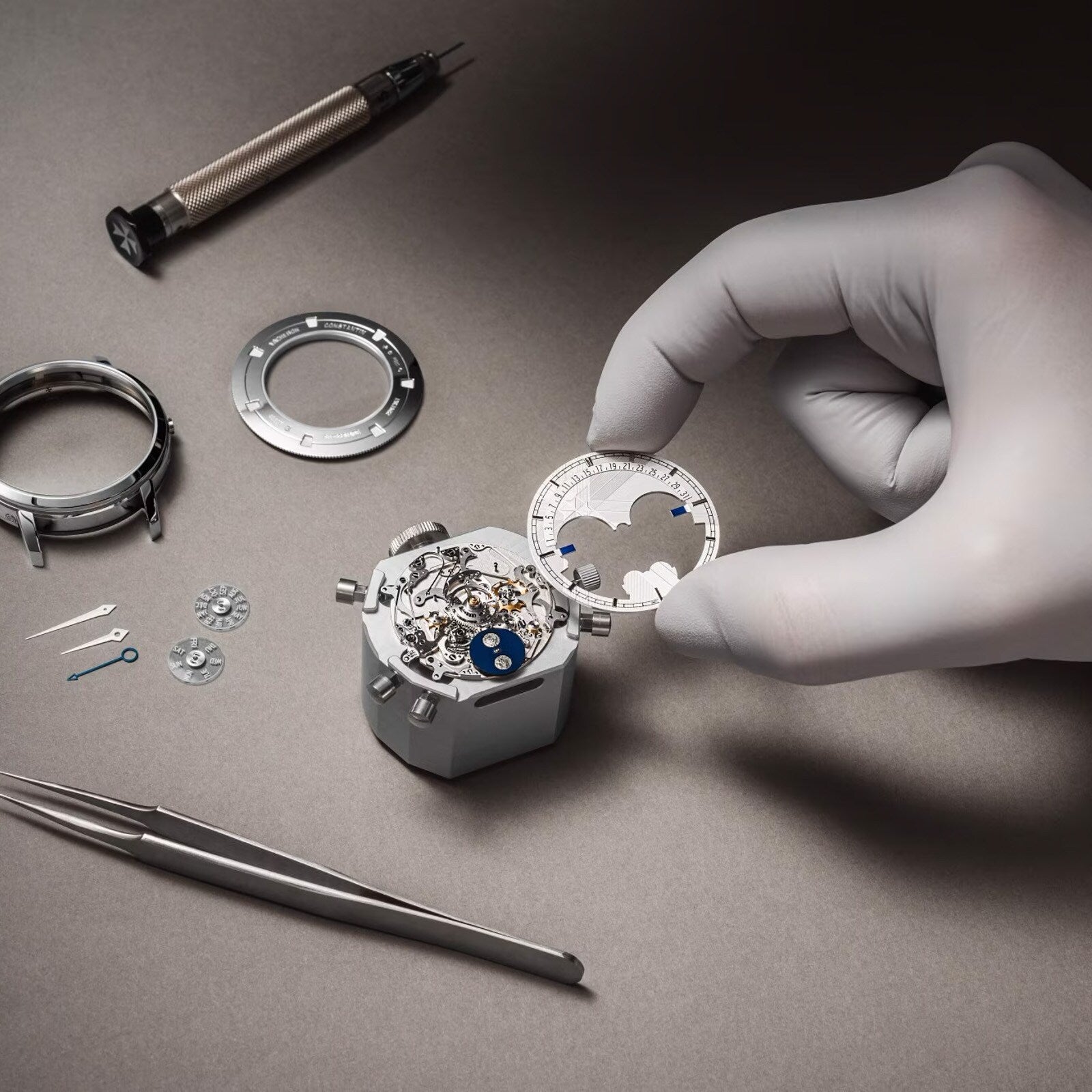 A gloved hand assembles a Vacheron Constantin Traditionnelle Perpetual Calendar Retrograde Date Openface movement on a work surface, surrounded by watch parts, a screwdriver, and tweezers.