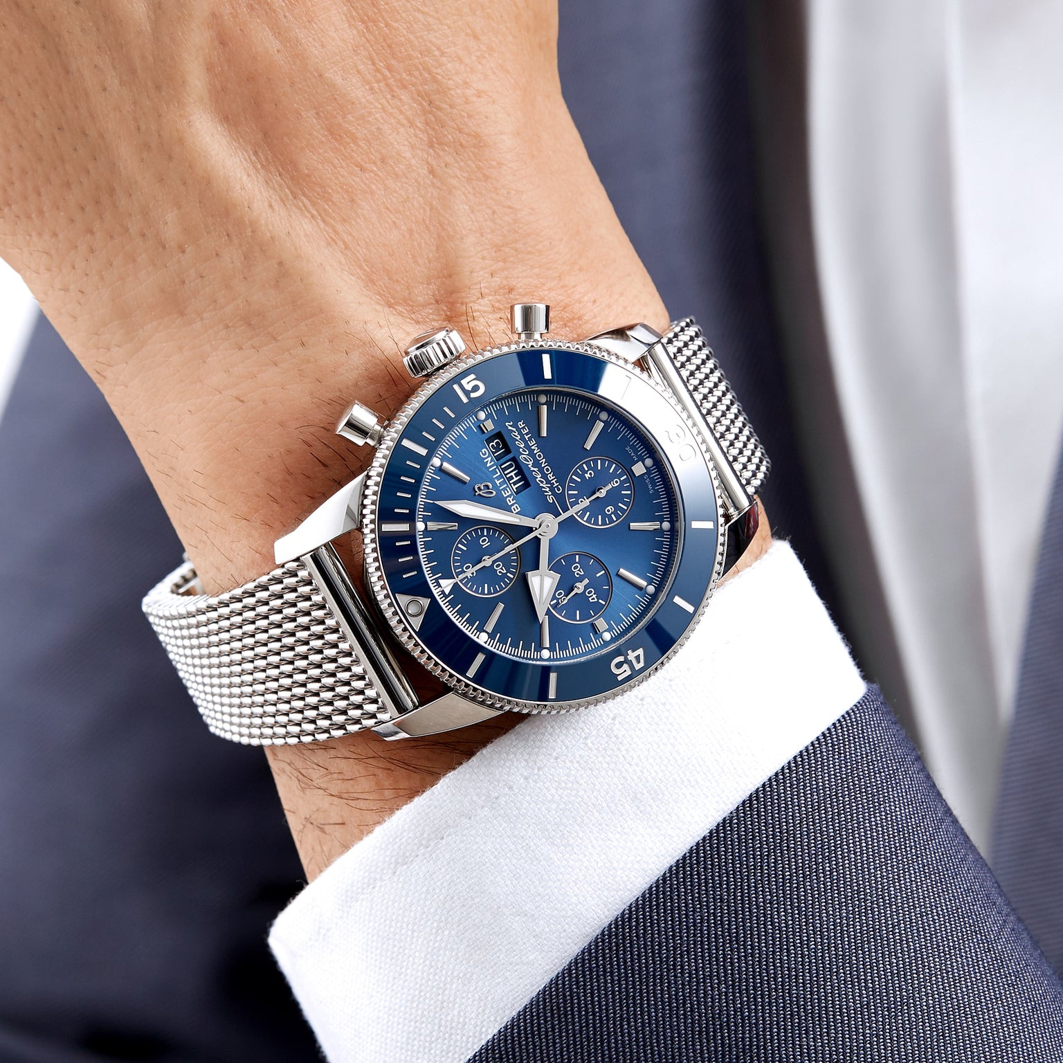 A person in a navy blue suit and white shirt wears the Breitling Superocean Heritage Chronograph 44 with a blue dial and stainless steel mesh bracelet.
