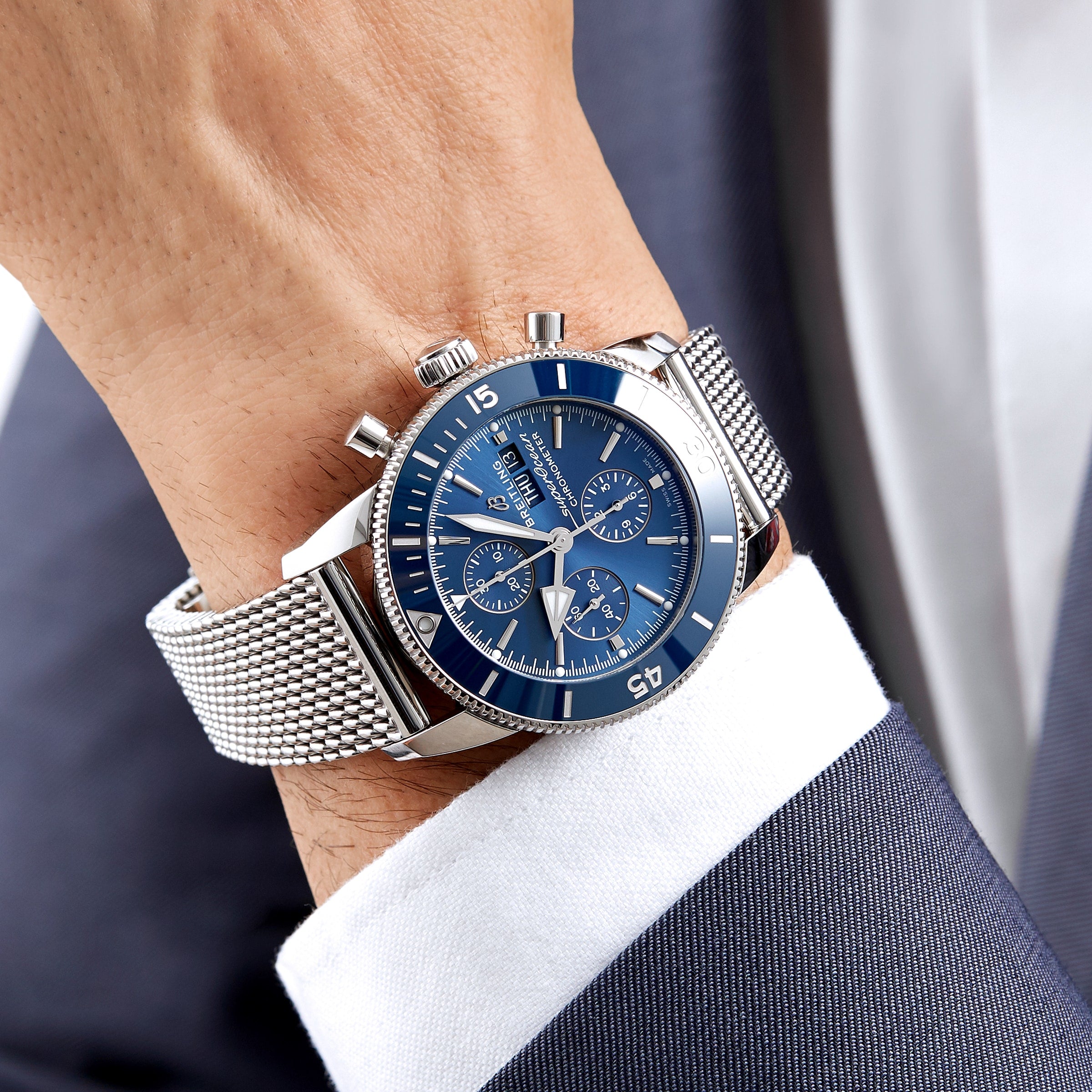 A person in a navy blue suit and white shirt wears the Breitling Superocean Heritage Chronograph 44 with a blue dial and stainless steel mesh bracelet.