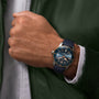 A person in a green jacket and white shirt showcases the Breitling Superocean Heritage B20 Automatic 42, featuring a blue dial, blue strap, silver casing, and notable water resistance.