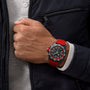 A person in a dark jacket and white shirt wears a Breitling Endurance Pro 44, a black analog wristwatch with red band and bezel, on their left wrist.