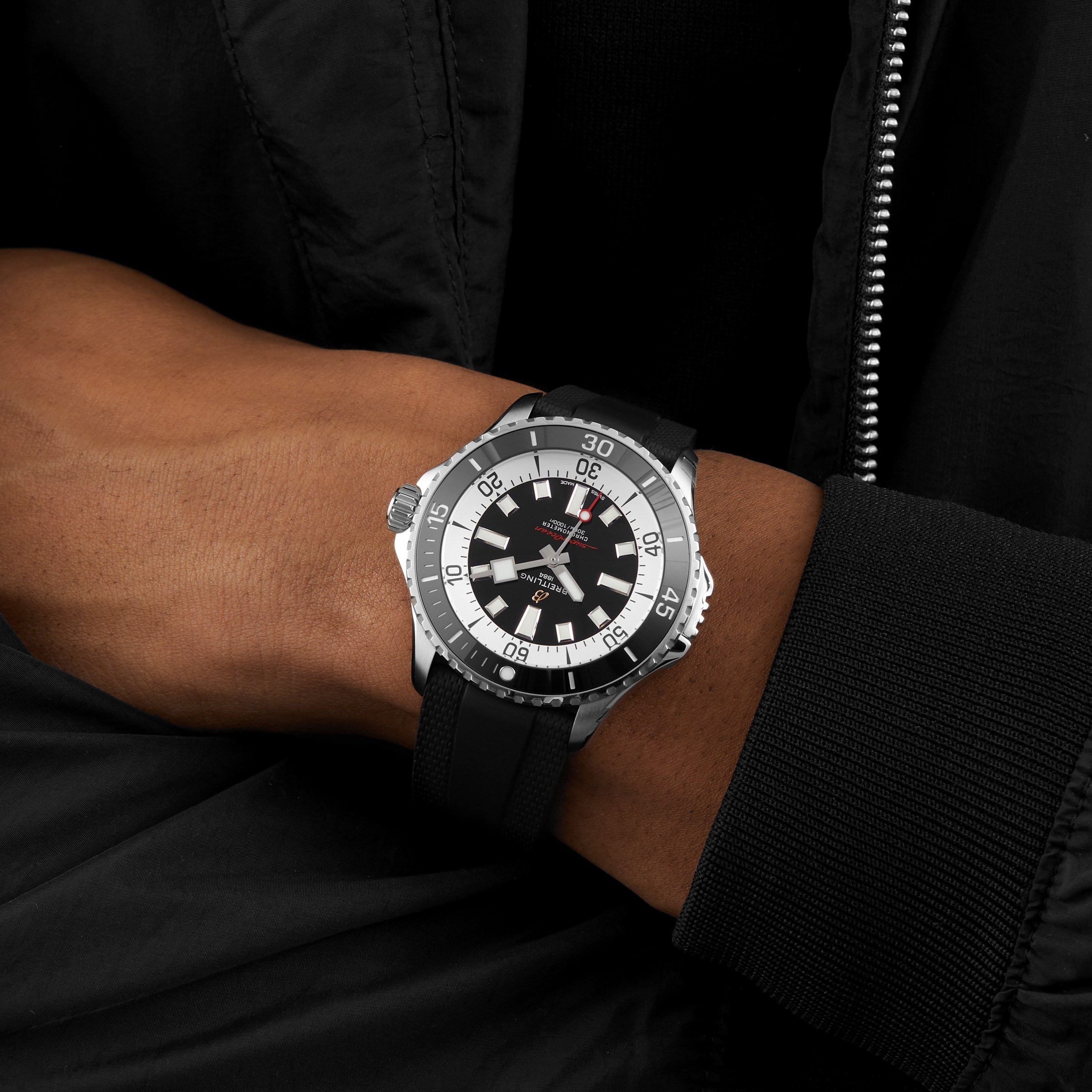 A person wearing a Breitling Superocean Automatic 46 black and silver wristwatch with a black strap and a black jacket.
