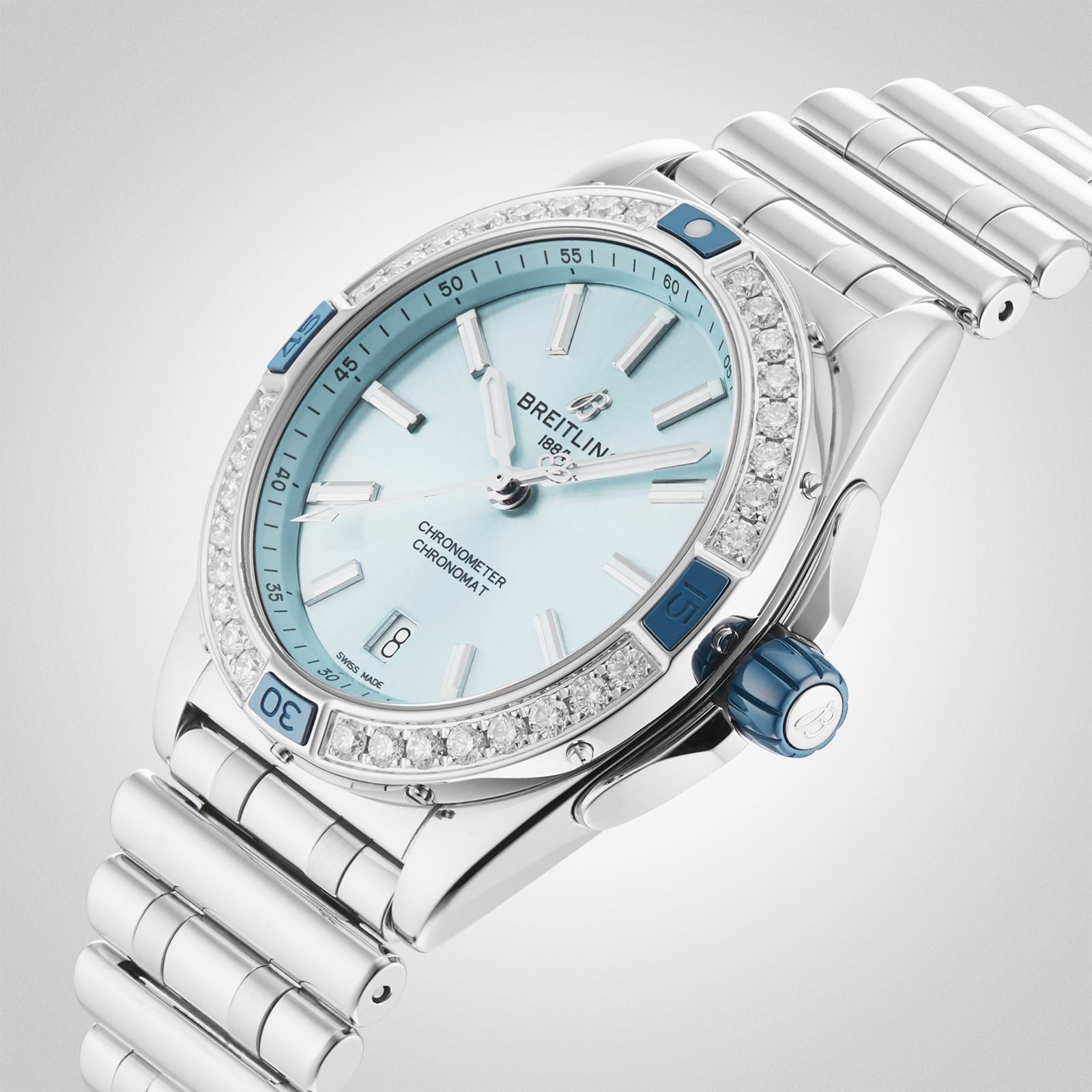 The Breitling Super Chronomat Automatic 38 features a silver case, light blue dial, diamond-studded bezel, date at 3 o’clock, metal bracelet, and reliable Breitling 17 calibre—all set on a light gradient background.