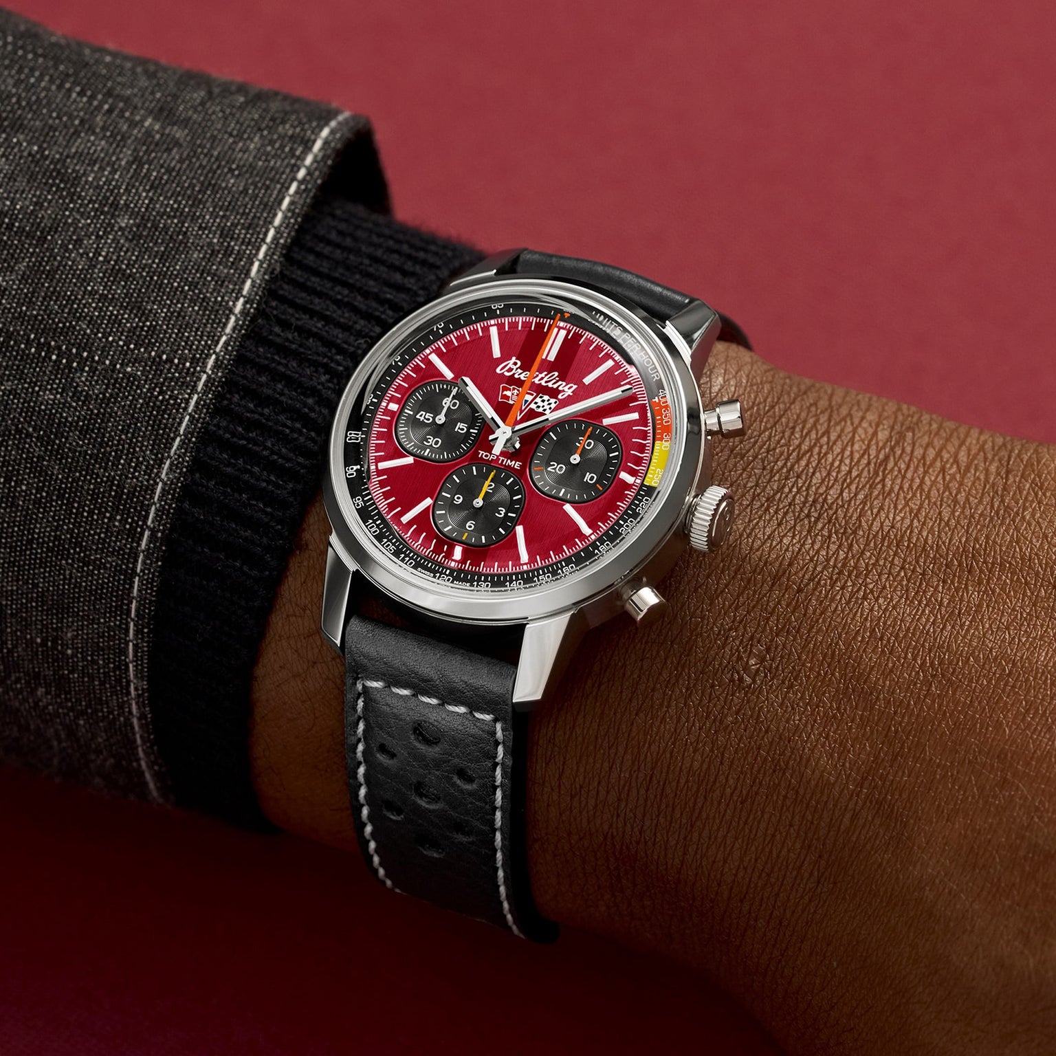 A person wearing the Breitling Top Time B01 Chevrolet Corvette watch—with a red dial, three subdials, black leather strap, and silver case—stands against a burgundy background.
