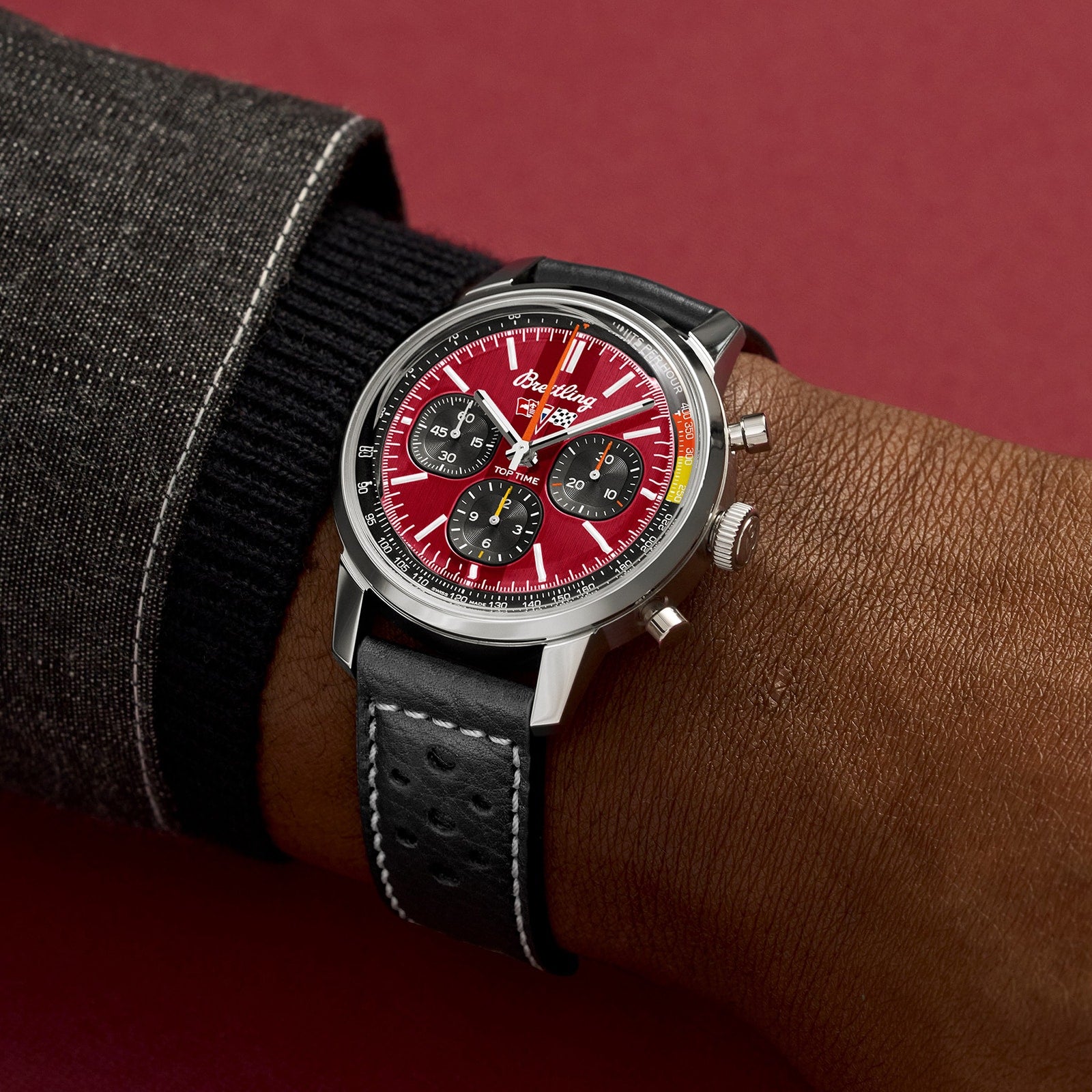 A person wearing the Breitling Top Time B01 Chevrolet Corvette watch—with a red dial, three subdials, black leather strap, and silver case—stands against a burgundy background.
