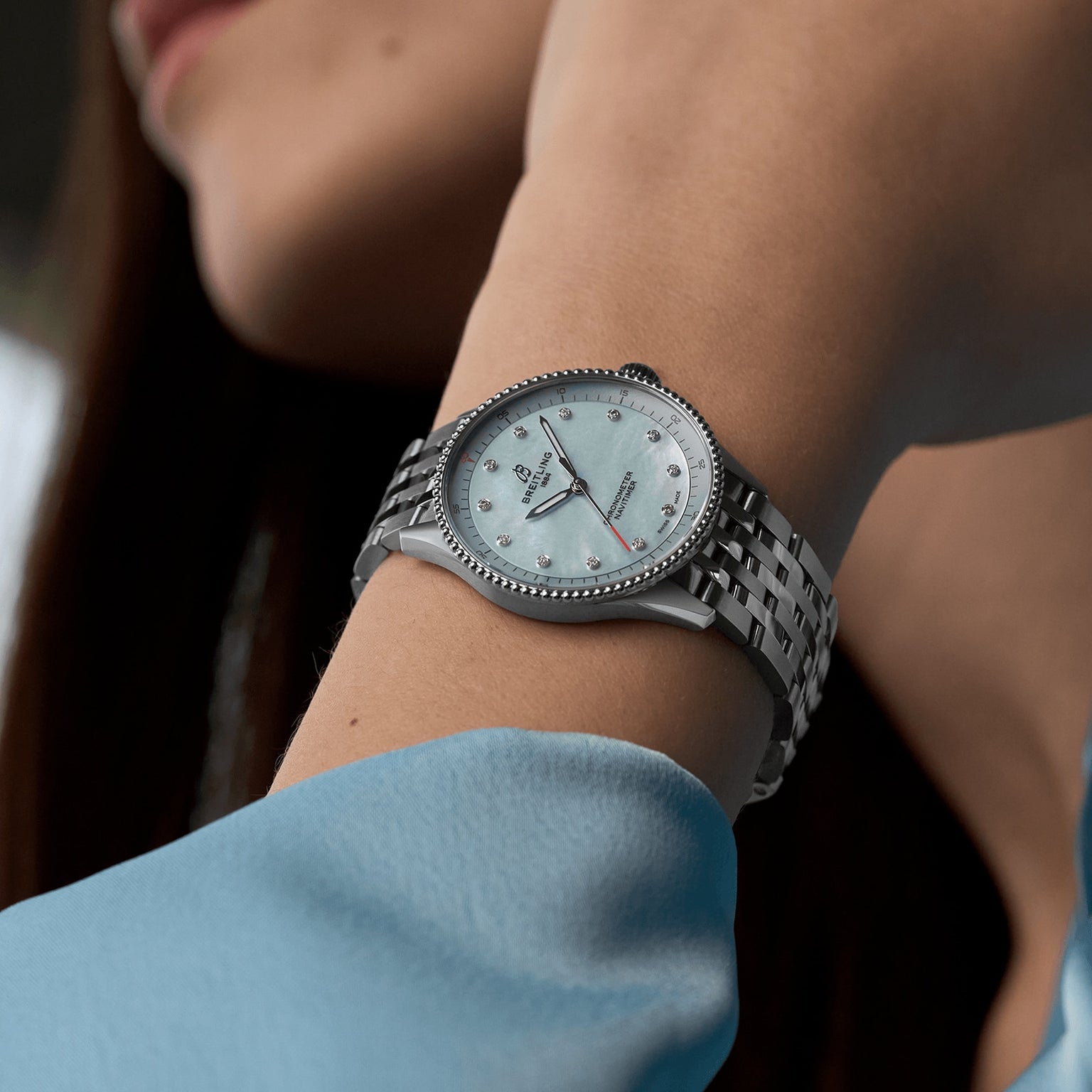 A person wearing a Breitling Navitimer Automatic GMT 41—a silver wristwatch with a white face and diamond hour markers, powered by the renowned Calibre 77 SuperQuartz™—dressed in a light blue long-sleeve garment.