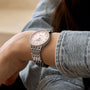 A person wearing the Breitling Navitimer 32, featuring a silver wristwatch with a pink face, diamond markers, and stainless steel case, dressed in a light denim long-sleeve shirt.