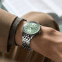 A person wearing a Breitling Navitimer Automatic 36 with a stainless steel case, mint green dial, and date display, paired with a beige jacket and grey sweater.